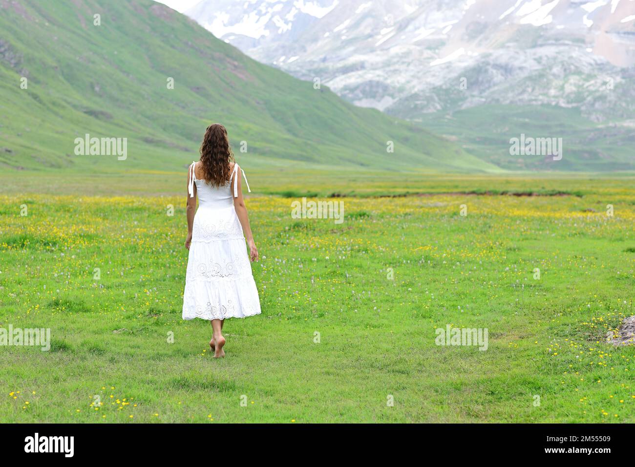 Back view of a woman in white dress walking in a green field in the mountain Stock Photo