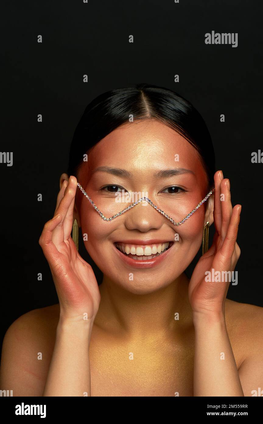 An Asian girl on a black background in the studio. Gold. Thin collarbones. The realm of beauty and perfume. Unusual glasses. Smile Stock Photo