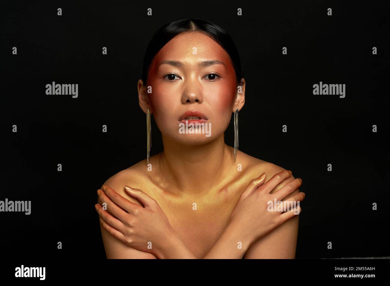 An Asian girl on a black background in the studio. Gold. Thin collarbones. Long gold earrings. The realm of beauty and perfume. Closed posture. Vulner Stock Photo