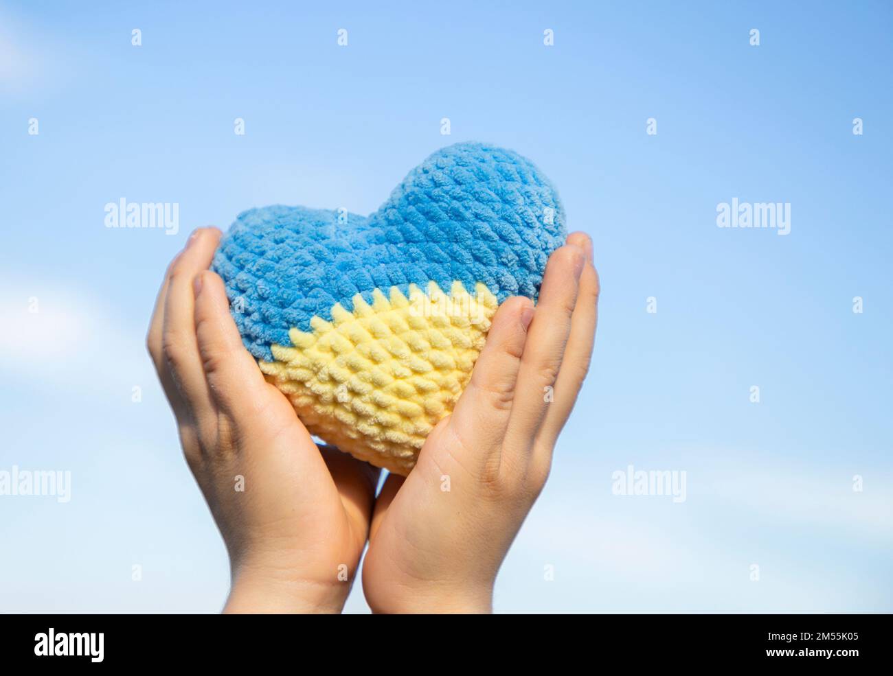 knitted heart of yellow and blue colors of Ukrainian flag in hands of child against sky. Symbol of love for Ukraine, patriotism. Support Ukraine. Stop Stock Photo