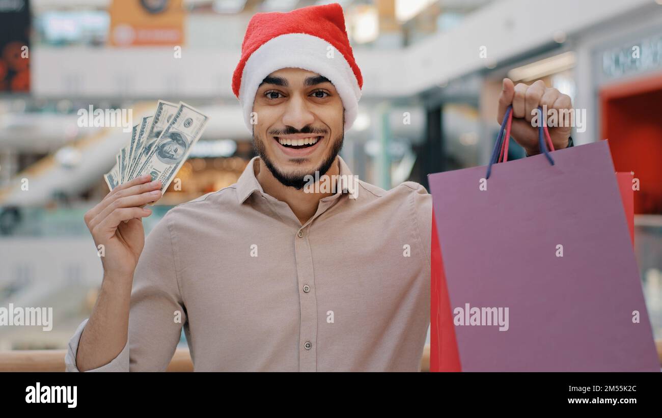 Indian happy Hispanic guy man in Santa X-mas Christmas hat in shopping mall looking camera showing holding money cash dollars and gift bags male Stock Photo