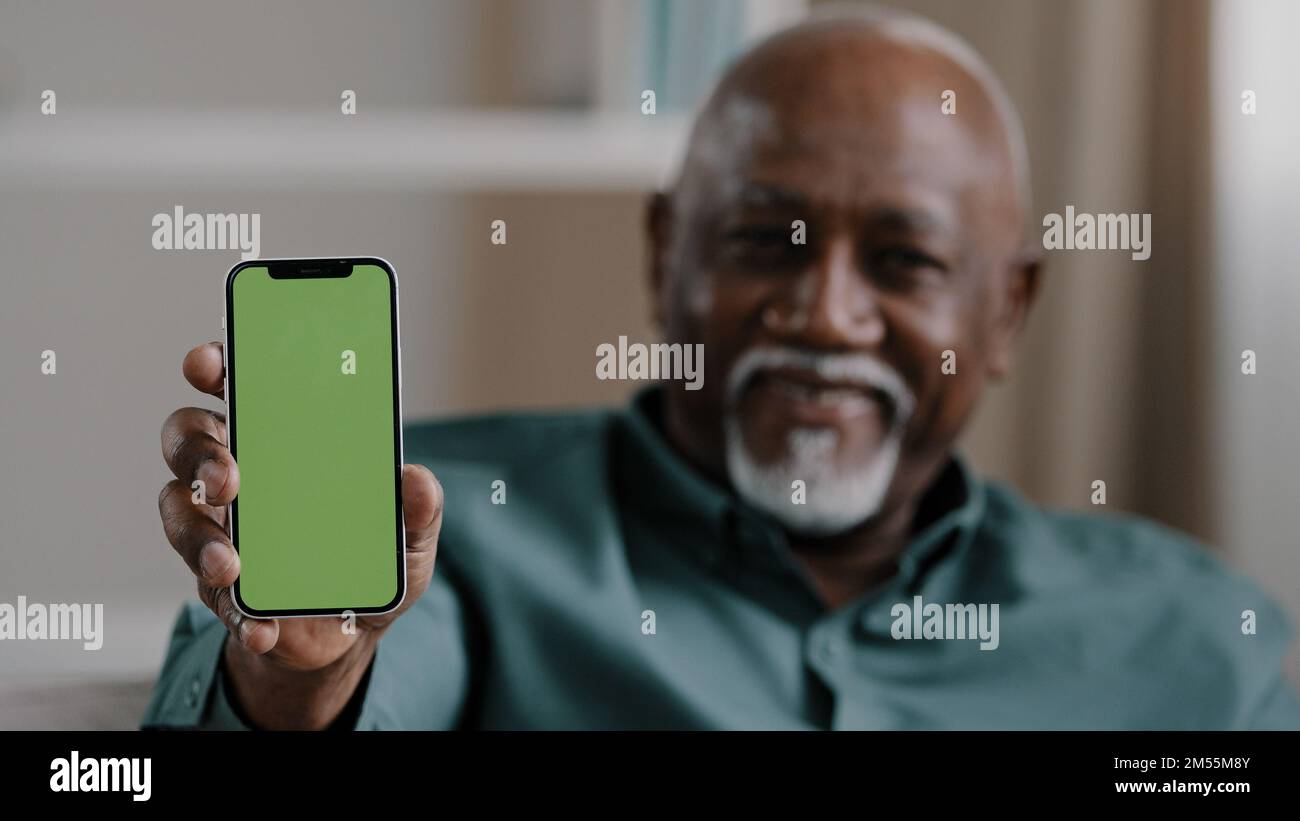 Old male African American biracial man at home sitting on couch businessman in office showing smartphone with empty green screen chromakey holding Stock Photo