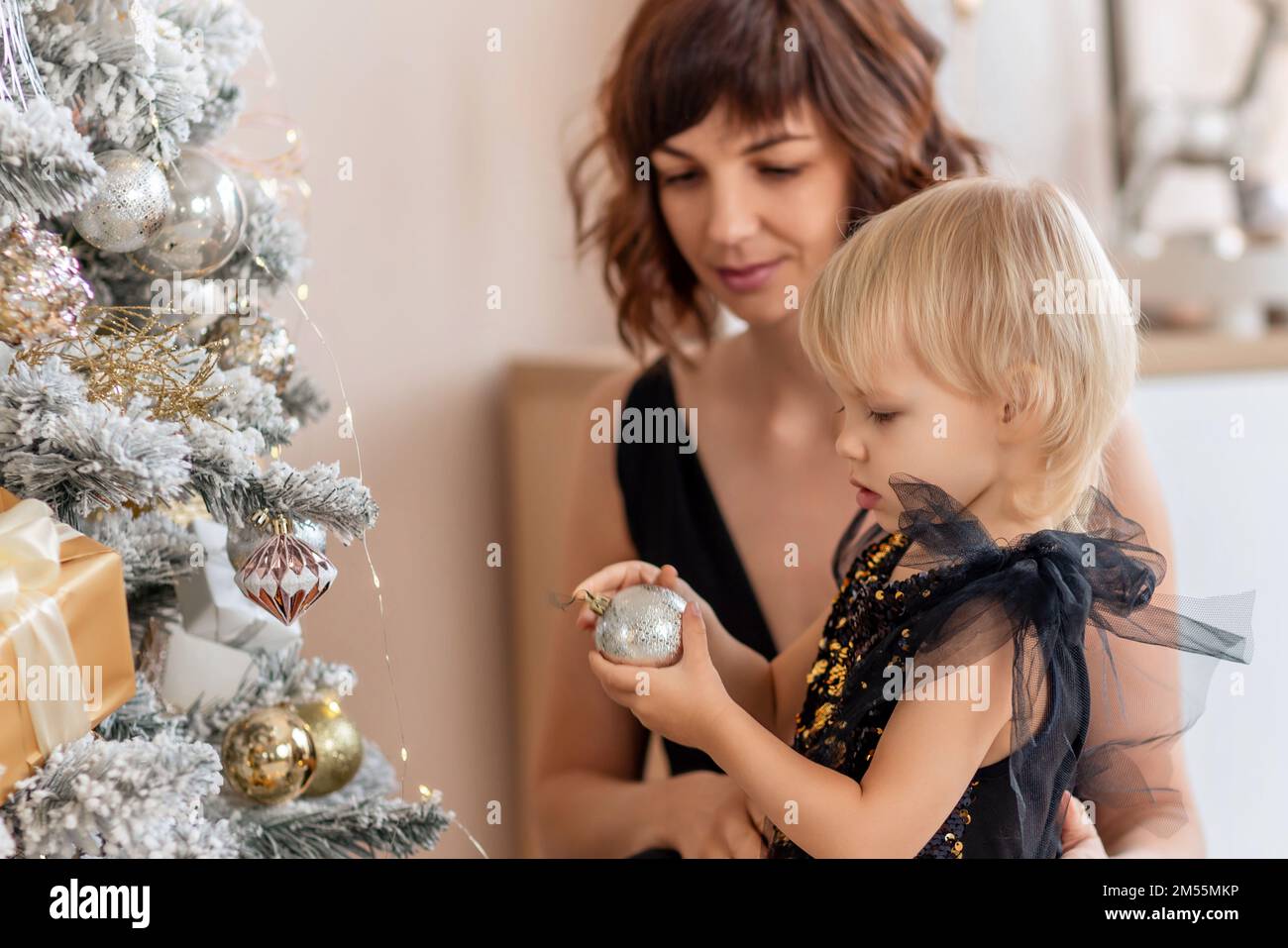 A mother with a 2 years old daughter decorates the Christmas tree. Both are dressed in black dresses, the daughter hangs a ball on the Christmas tree. Stock Photo