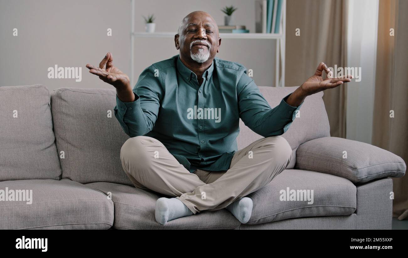 Happy elderly african american man sit on sofa in lotus position with folded fingers grandpa is meditating finds harmony relieves stress positive calm Stock Photo