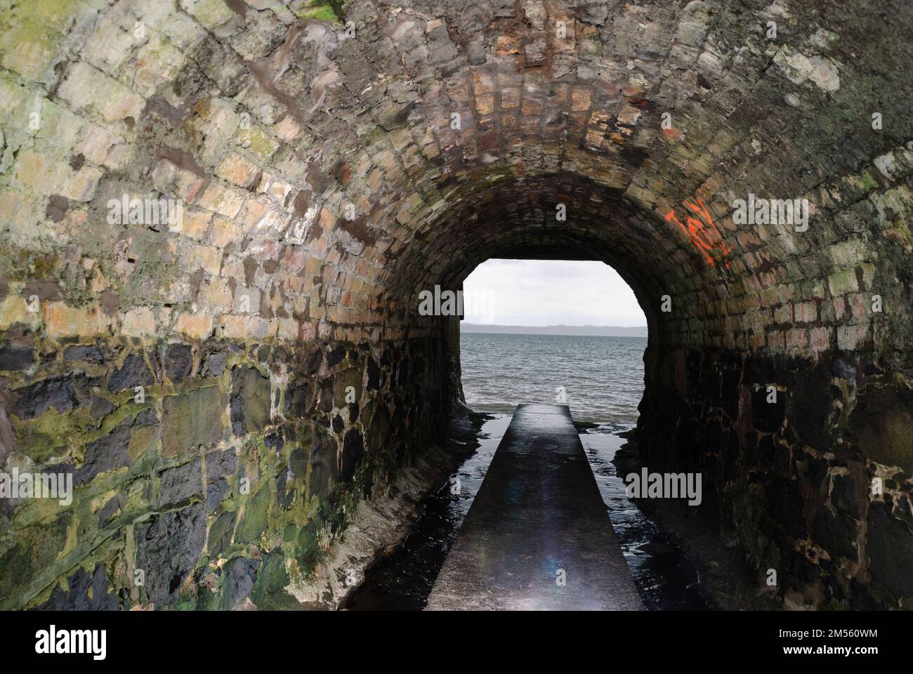 Dirty water runs down the sides of a path through a sewer tunnel to flow out to the sea. Stock Photo