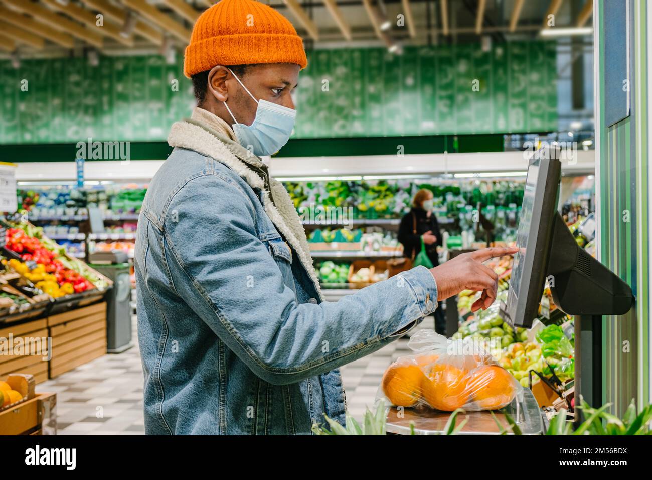Stylish African-American guy in warm jacket with disposable mask weighs fresh oranges with digital scale in light supermarket Stock Photo