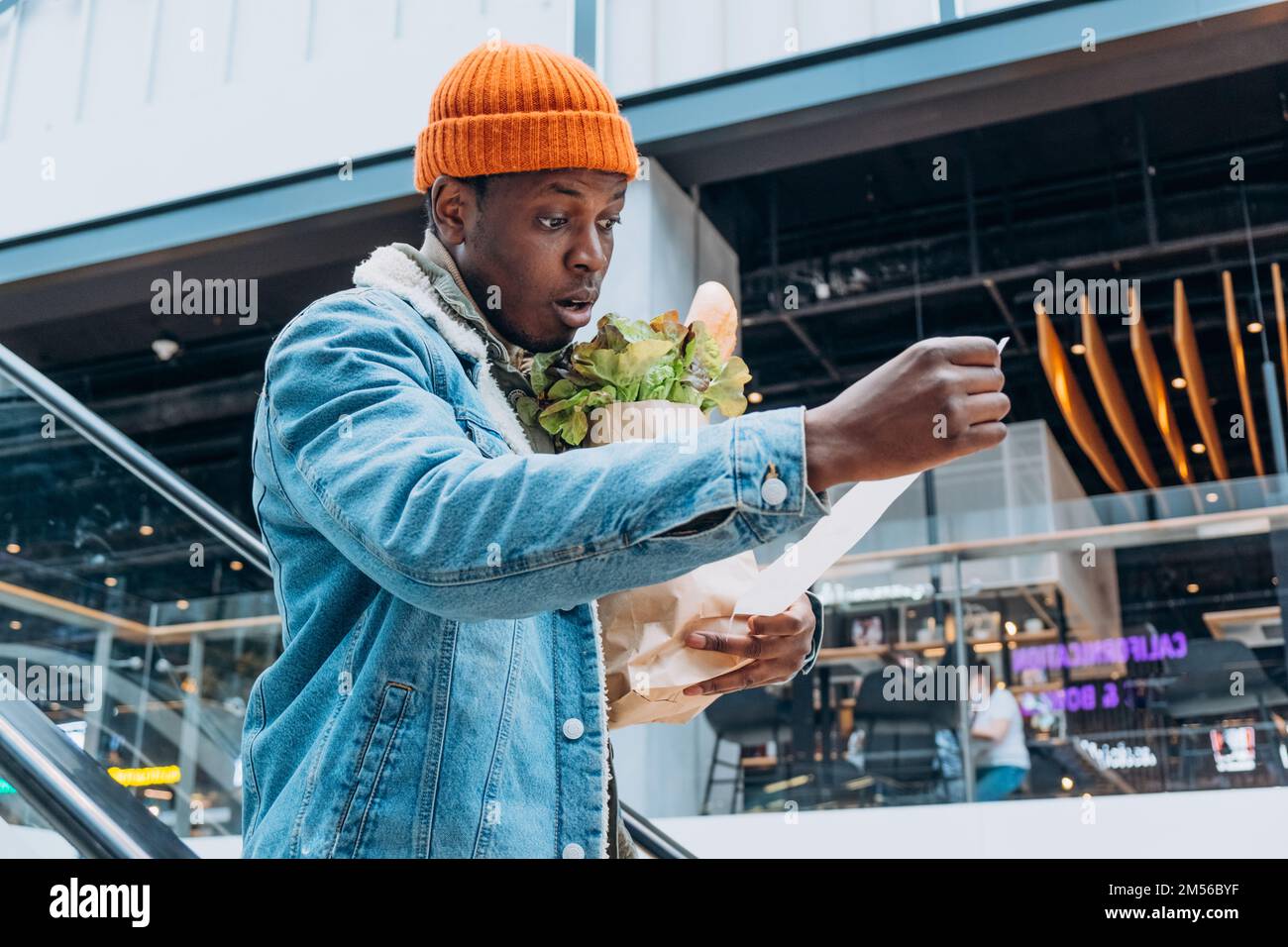 Doubting African-American person in denim jacket looks at sales paper receipt total holding pack with food products on escalator Stock Photo