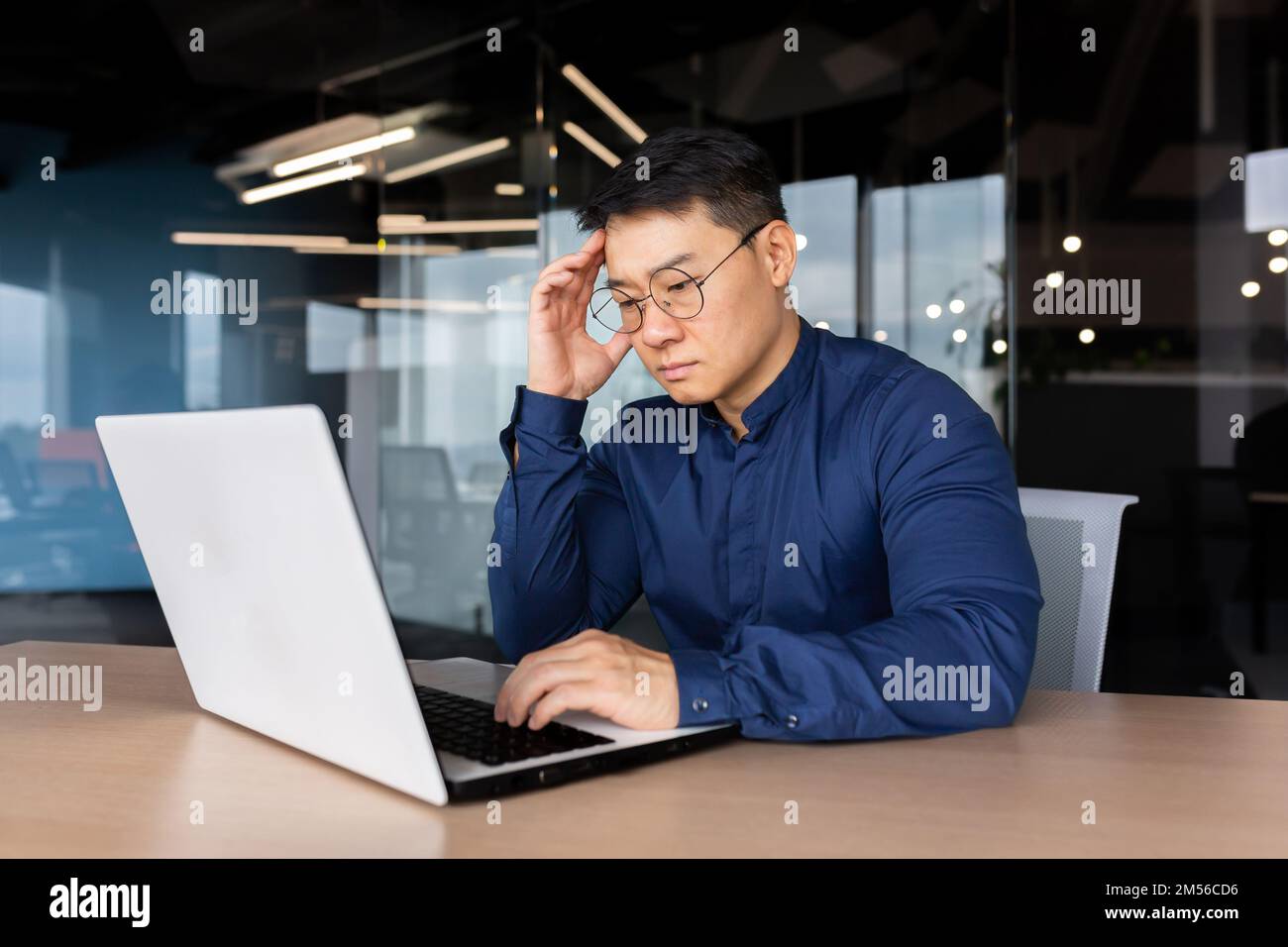 Thoughtful and serious male Asian student sits in the office at the table, works on a laptop. Passes the exam, worries, studies online through a video call. He holds his head with his hand. Stock Photo