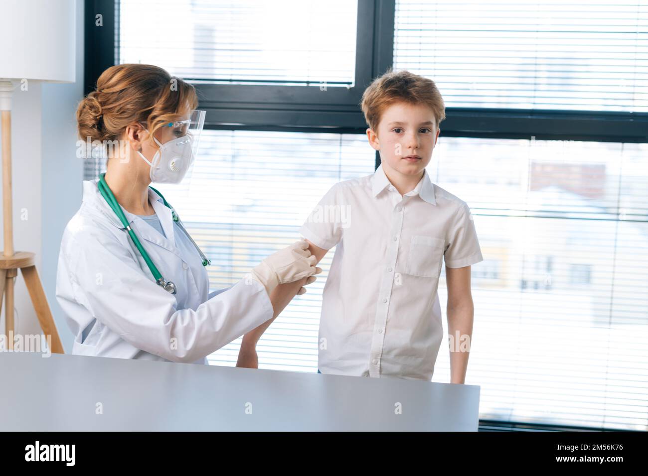 Portrait of female doctor wearing white uniform applying plaster on shoulder of child boy after vaccination injection by window. Stock Photo