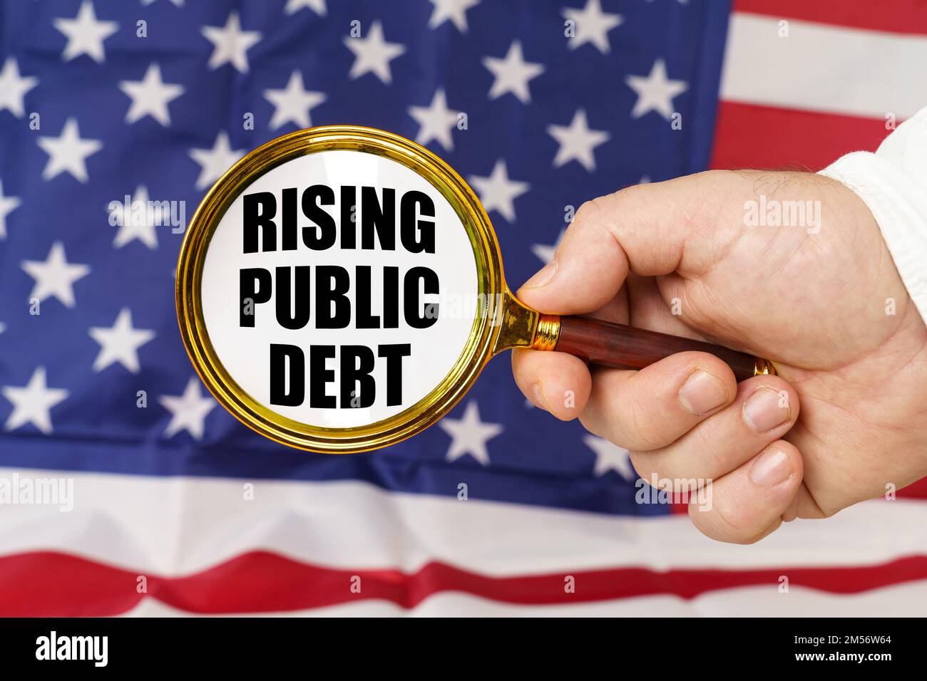 In front of the American flag, a man holds a magnifying glass in his hand with the inscription - rising public debt. Stock Photo