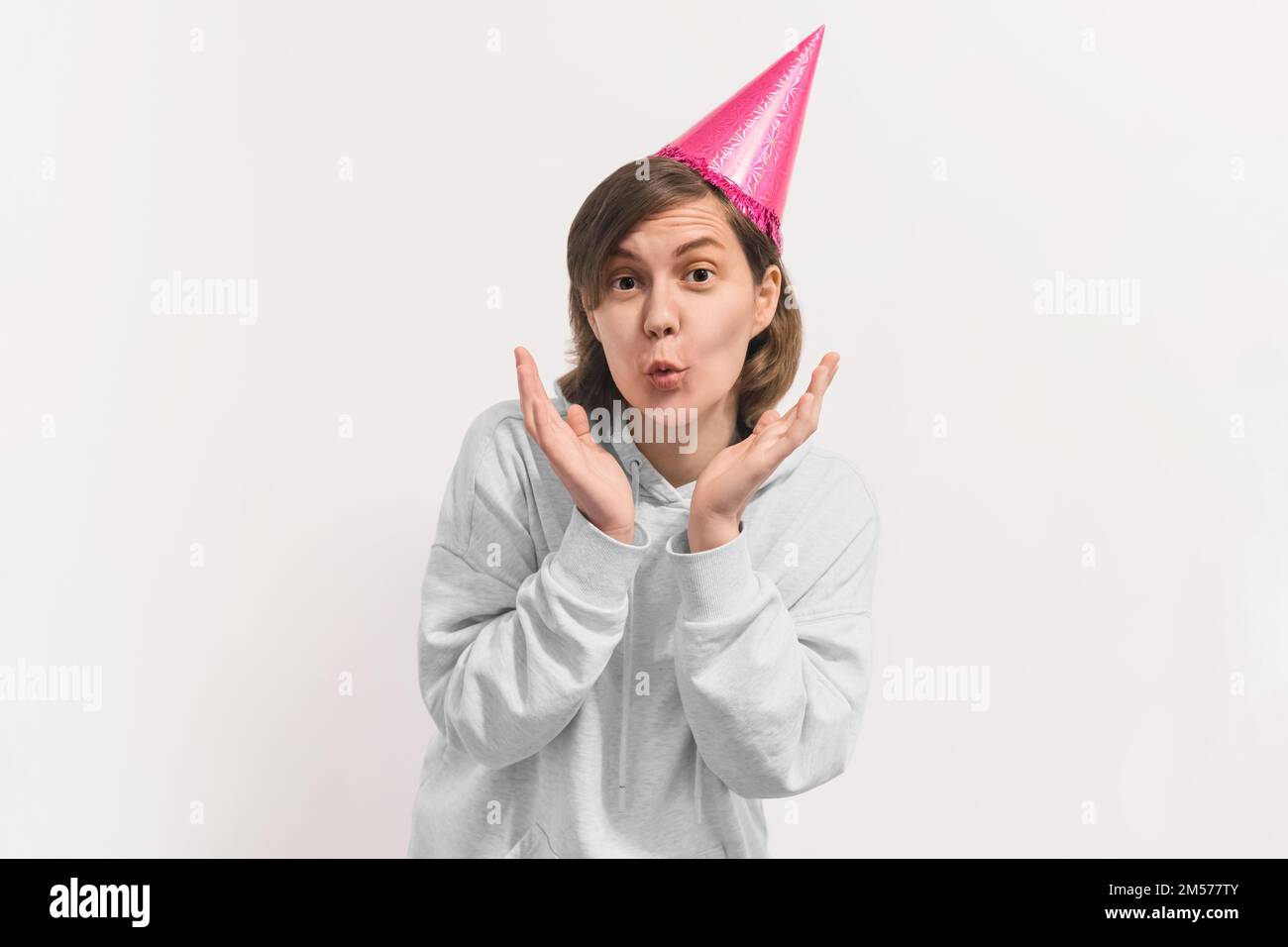 Young surprised woman in pink birthday hat isolated on white background.  Excited girl in hoodie holding her hands near face. Lifestyle concept Stock Photo