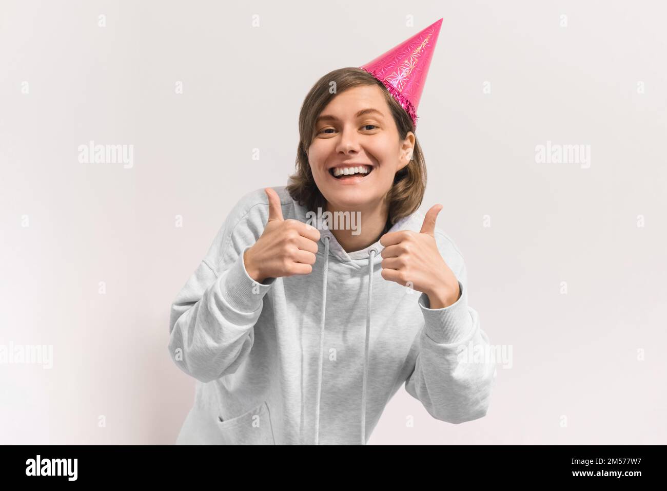 Young pretty woman in birthday hat laughing and rising her hands in Super gesture. Studio portrait of cheerful girl isolated on white background Stock Photo