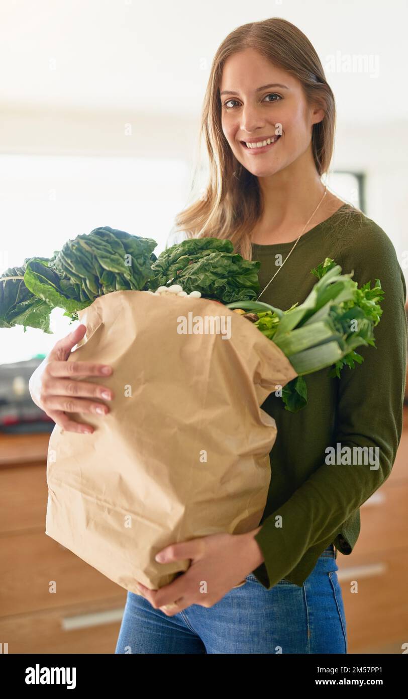 I only buy the freshest produce. Portrait of a smiling young woman standing in her kitchen carrying a paper bag full of groceries. Stock Photo