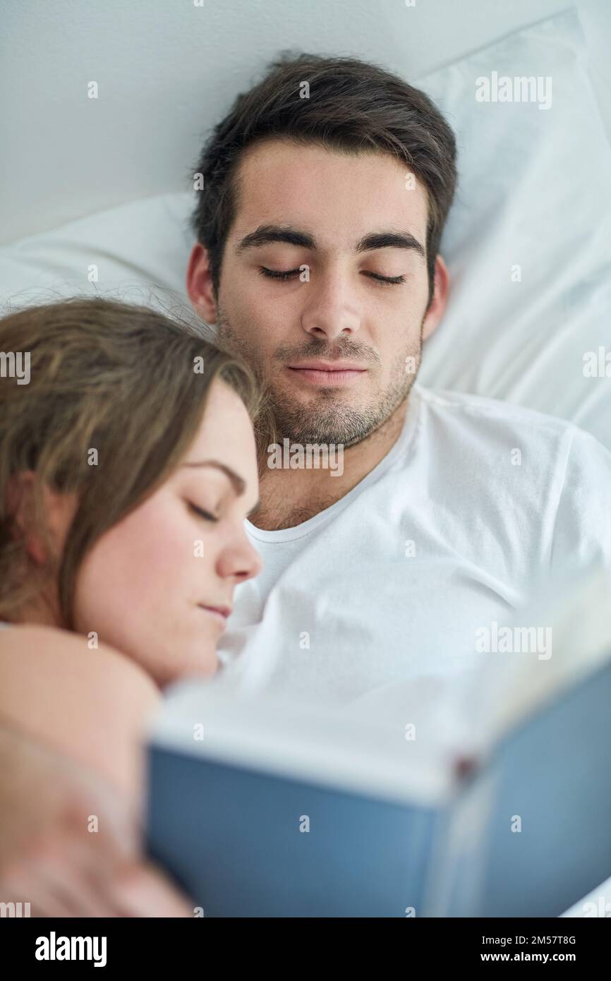 Enjoy the bliss of homeliness. a young couple reading a book in bed while his wife sleeps on his chest at home. Stock Photo