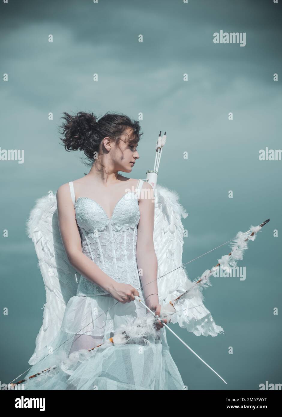 Valentin teenager girl with bow arrow ready to shoot. Angel teen in white angels dress with feathers wings on sky. Valentines Day. Stock Photo