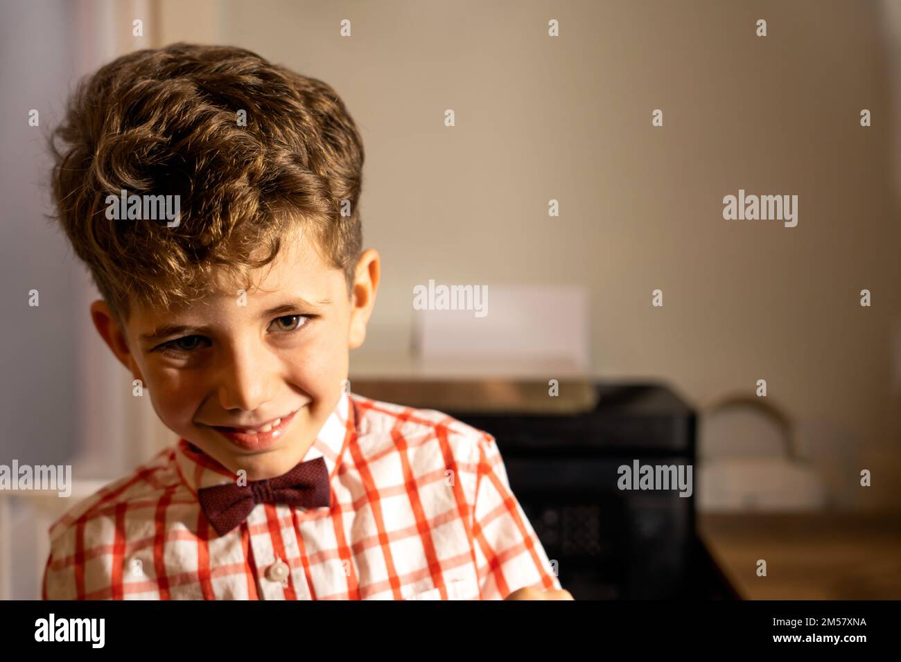 Cute preteen boy with curly brown hair in checkered shirt and bow tie in light room looking at camera Stock Photo