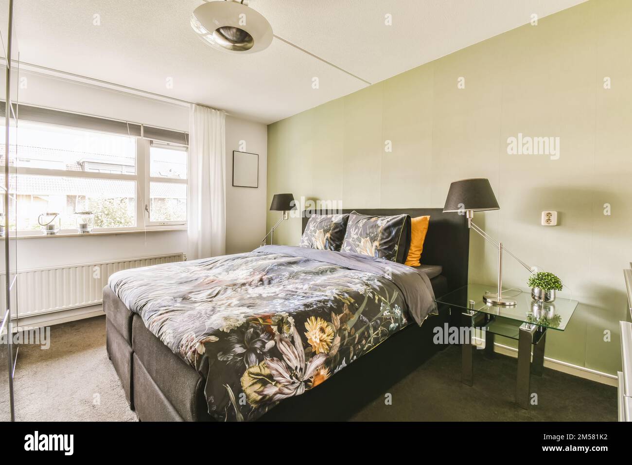 a bedroom with a bed, desk and lamp on the side table in front of the window looking out onto the street Stock Photo