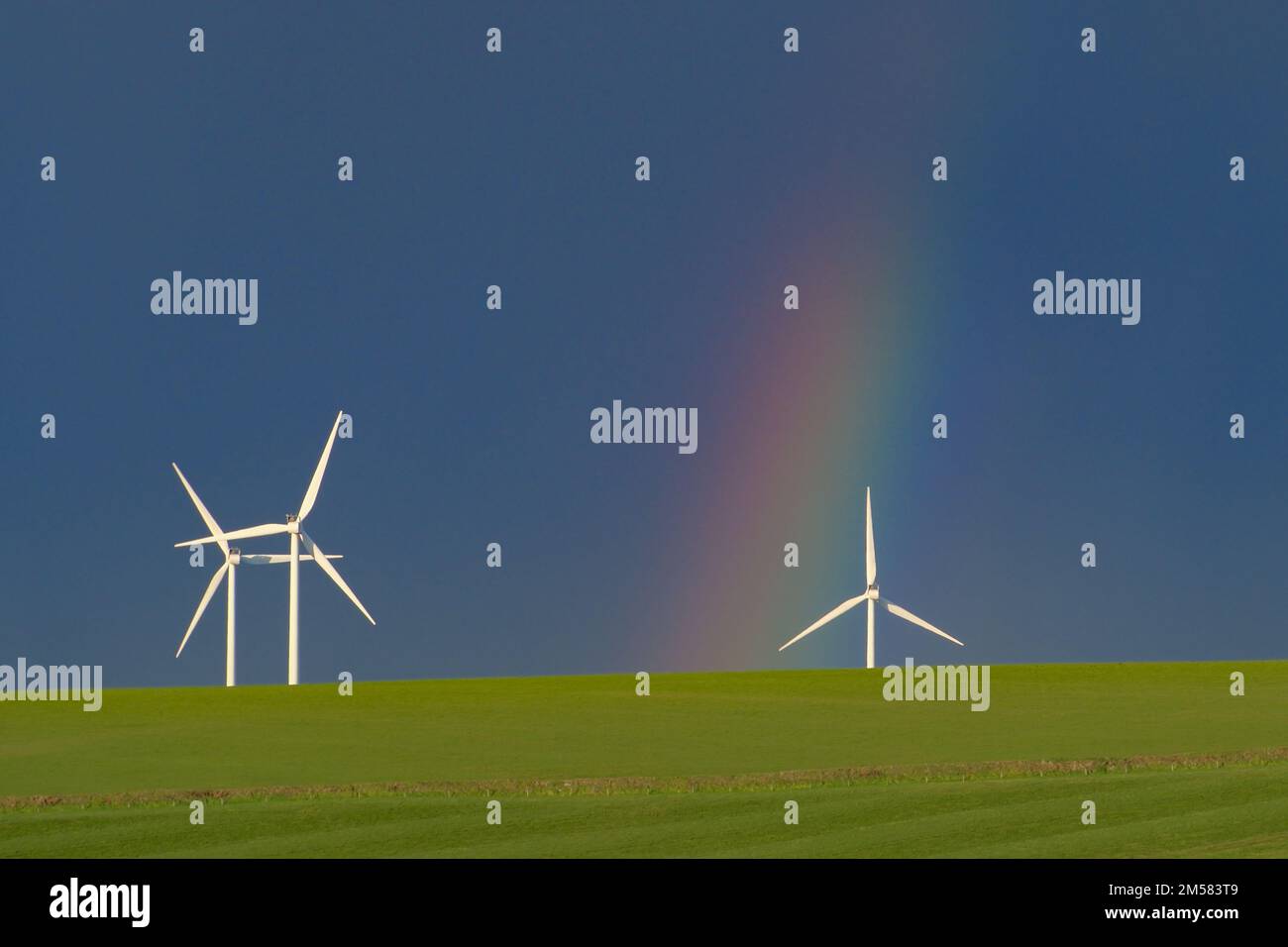 Wind turbines on a green field with a rainbow touching ground behind one of the turbines Stock Photo