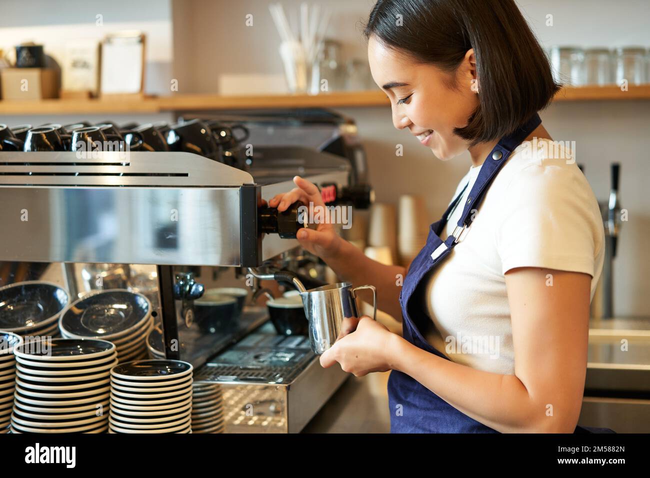 Smiling young asian barista girl, using coffee machine steamer
