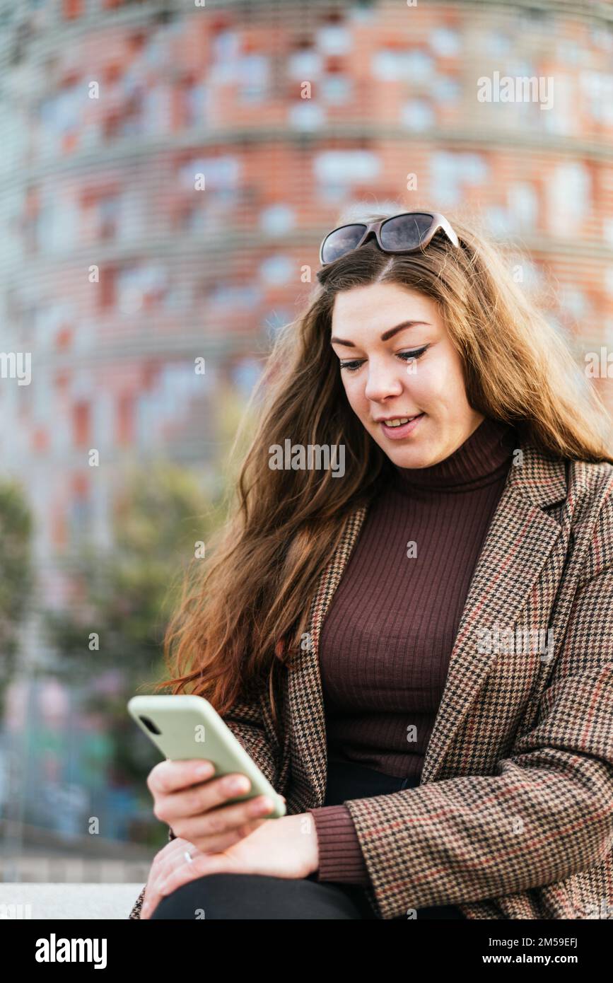 Positive female in outerwear with long clothes smiling and using mobile phone while resting on blurred background of city street. Stock Photo