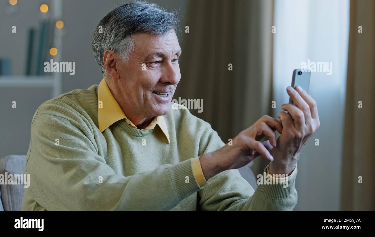 Happy elderly senior adult man holding phone make video call smiling grandfather looking at smartphone screen remote communicate use modern device Stock Photo