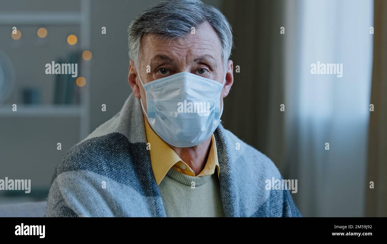 Close-up adult mature man wearing medical mask looking at camera sick old male sitting indoors in blanket portrait serious elderly retired pandemic Stock Photo