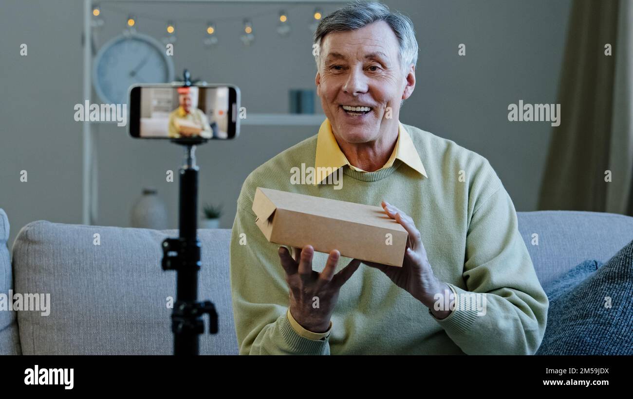 Elderly vlogger record videoblog online stream on social networks old man conducts live broadcast grandfather unpacking mystery box making video on Stock Photo