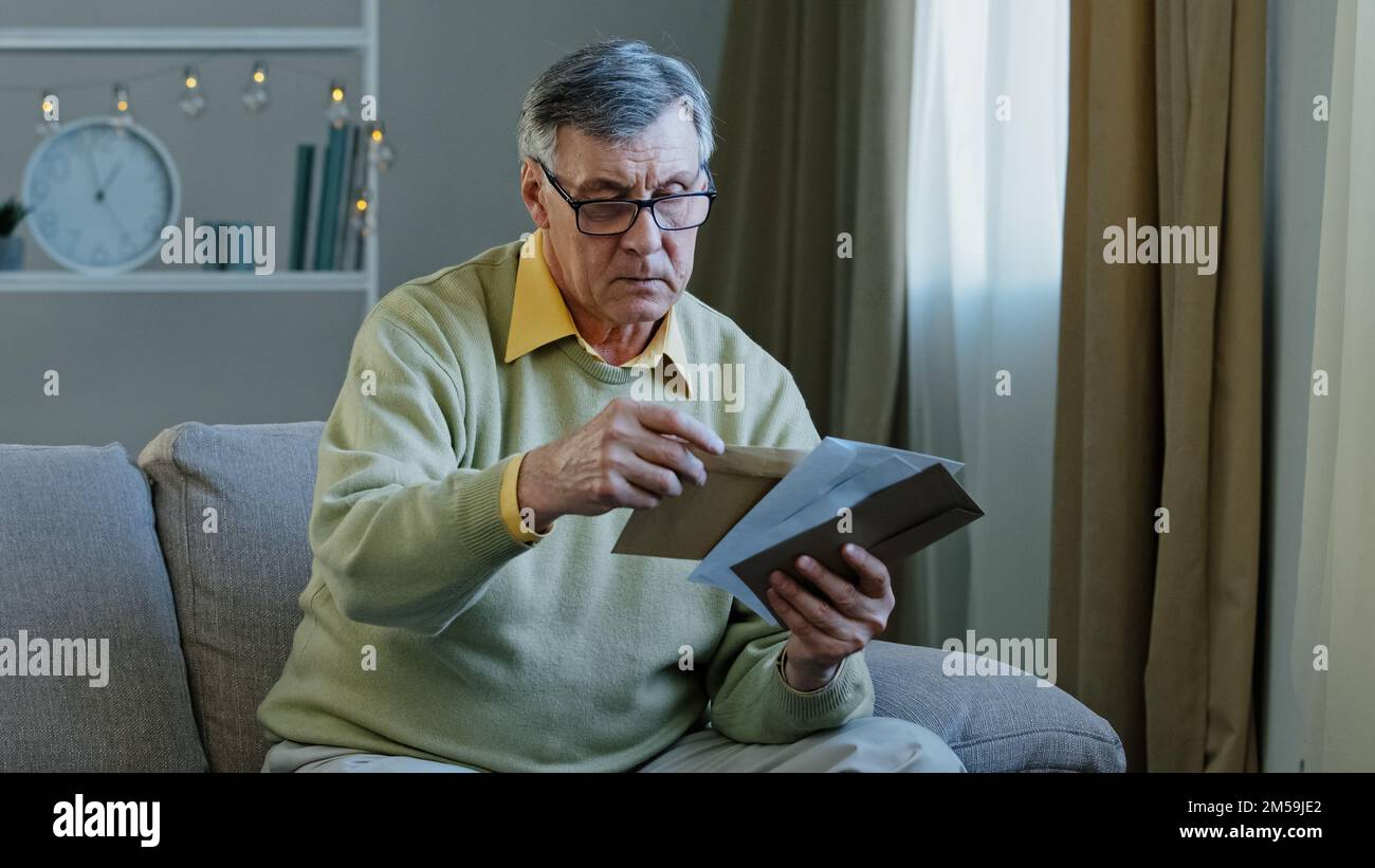 60s elderly man recipient with glasses sorting letters in living room concentrated mature grandfather sitting on sofa looking at correspondence notice Stock Photo