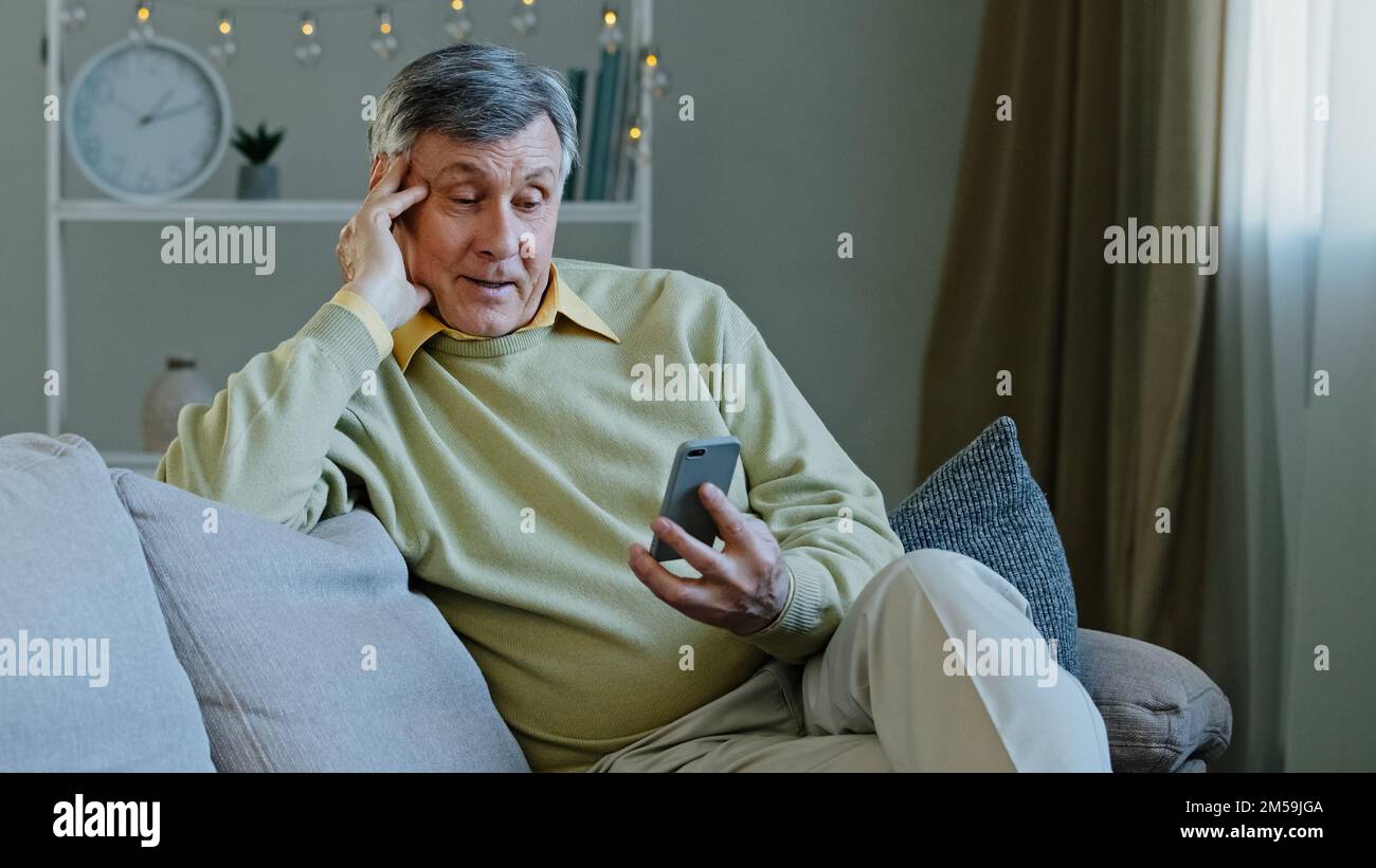 Happy elderly adult man sitting on sofa hold gadget studying app on smartphone makes online order smiling grandfather resting in room reading news Stock Photo