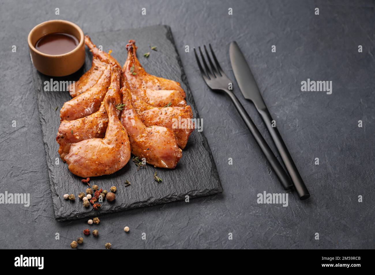 Delicacies. Grilled BBQ quail legs in marinade and spices on black slate with  sauce on a dark background. Healthy diet food. Copy space. Stock Photo