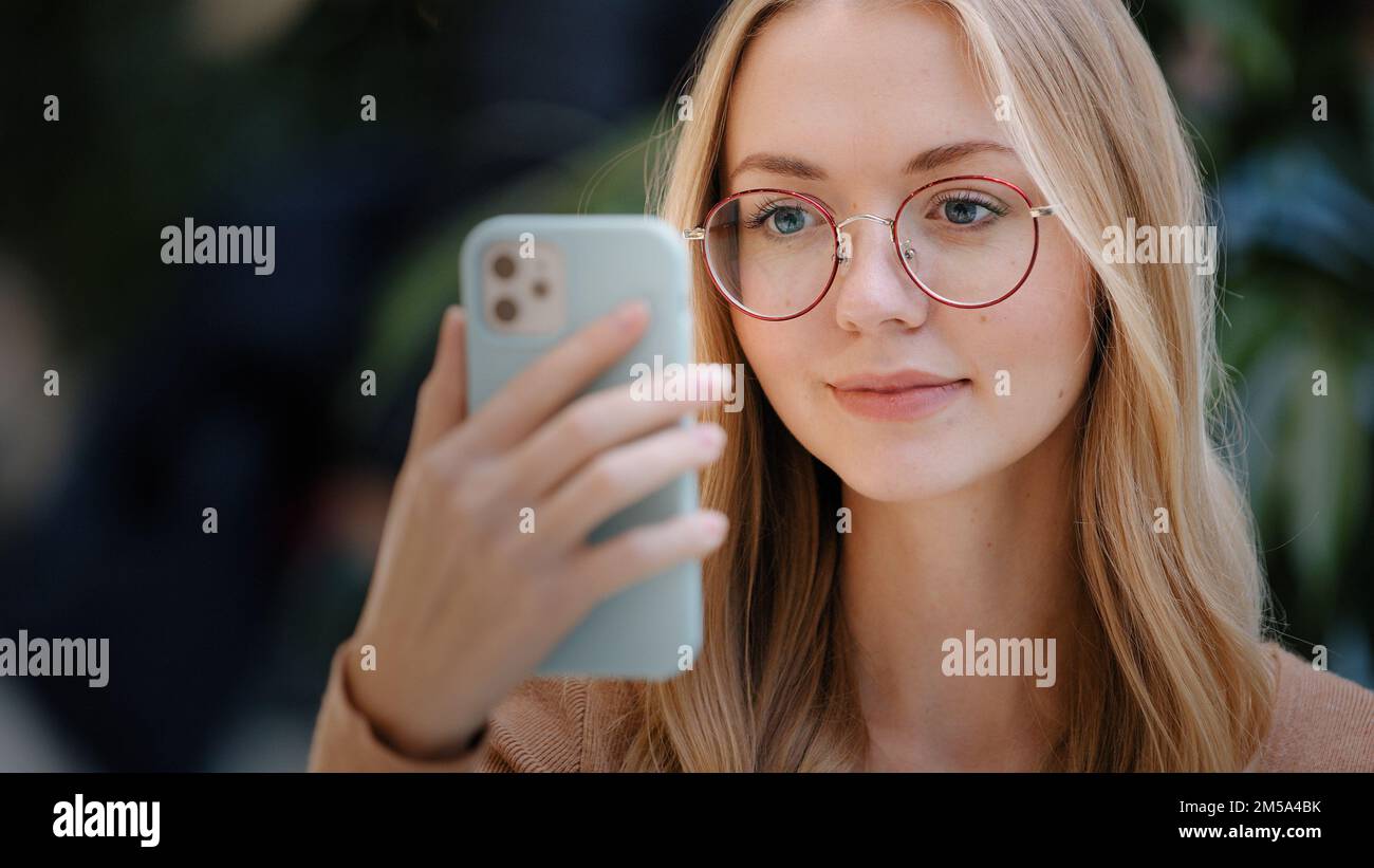 Portrait Caucasian girl blonde woman lady female in glasses looks at smartphone screen of mobile phone online message with offer reading news watching Stock Photo
