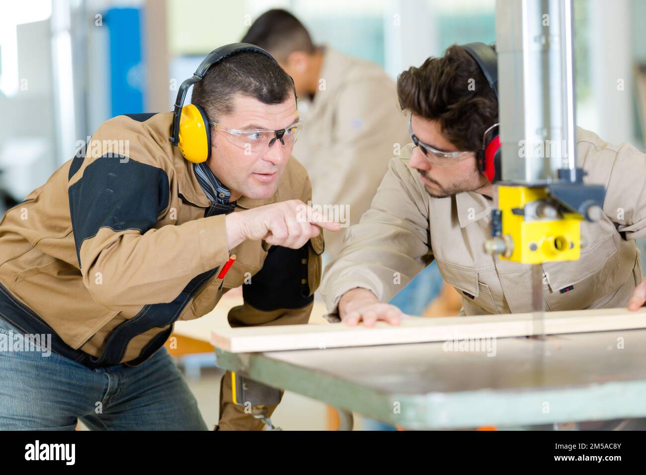 experienced carpenter guiding apprentice using bench saw Stock Photo