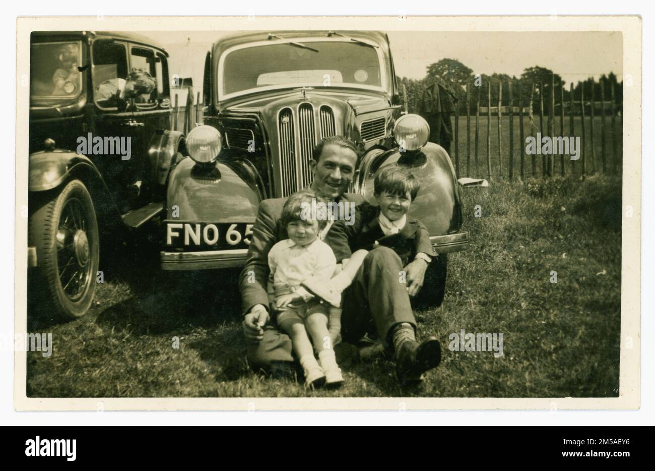 Original charming late 1930's era postcard of two young children sitting in front of a Ford Anglia on a day out with their dad, maybe a rally. Canterbury area, Kent England, UK. Stock Photo