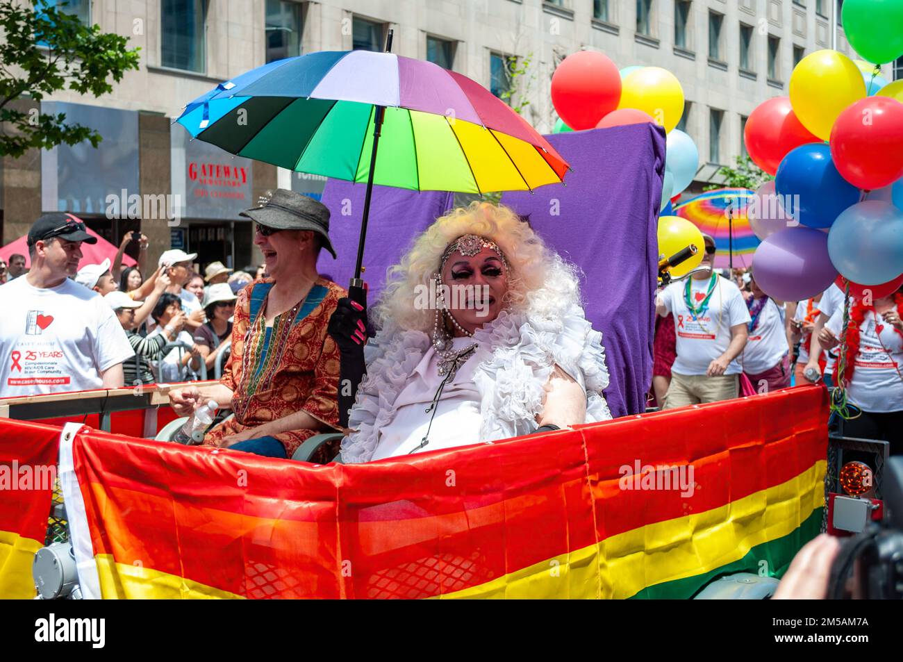 Two people in a small float covered in rainbow symbols partake in the LGBTQ+ celebration event. One of them is dressed as a drag queen. Stock Photo