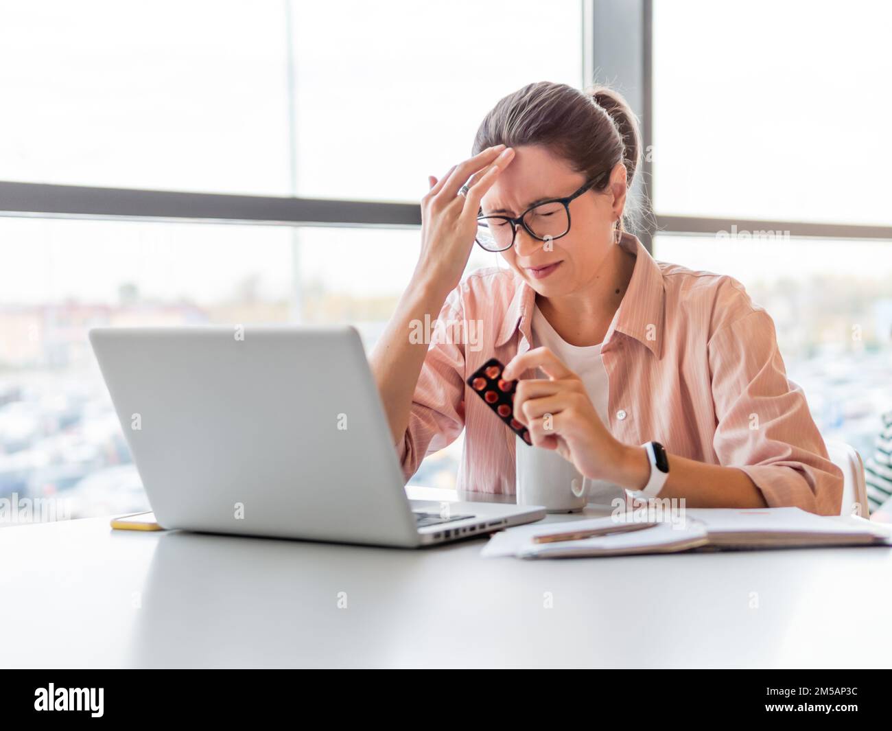 Frowning woman looks on medicine pills while working with laptop. Mental health problems, emotional burnout or headache. Modern office at co-working c Stock Photo