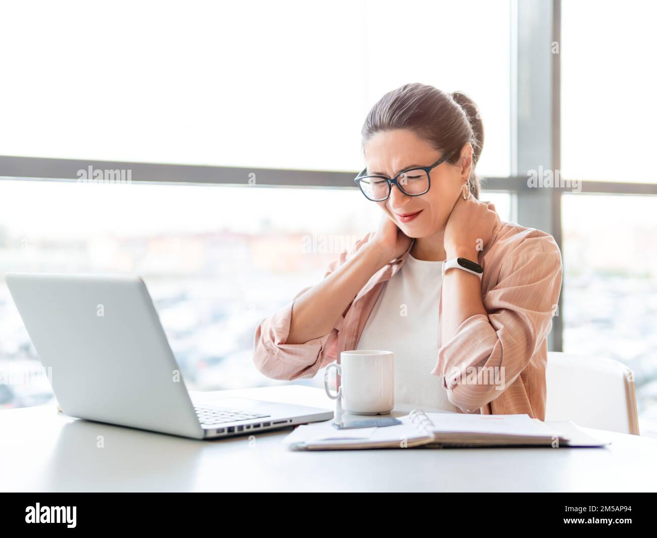 Woman winces from pain in her neck. Female office employee tries to stretch stiff neck muscles while working with laptop. Modern office at co-working Stock Photo