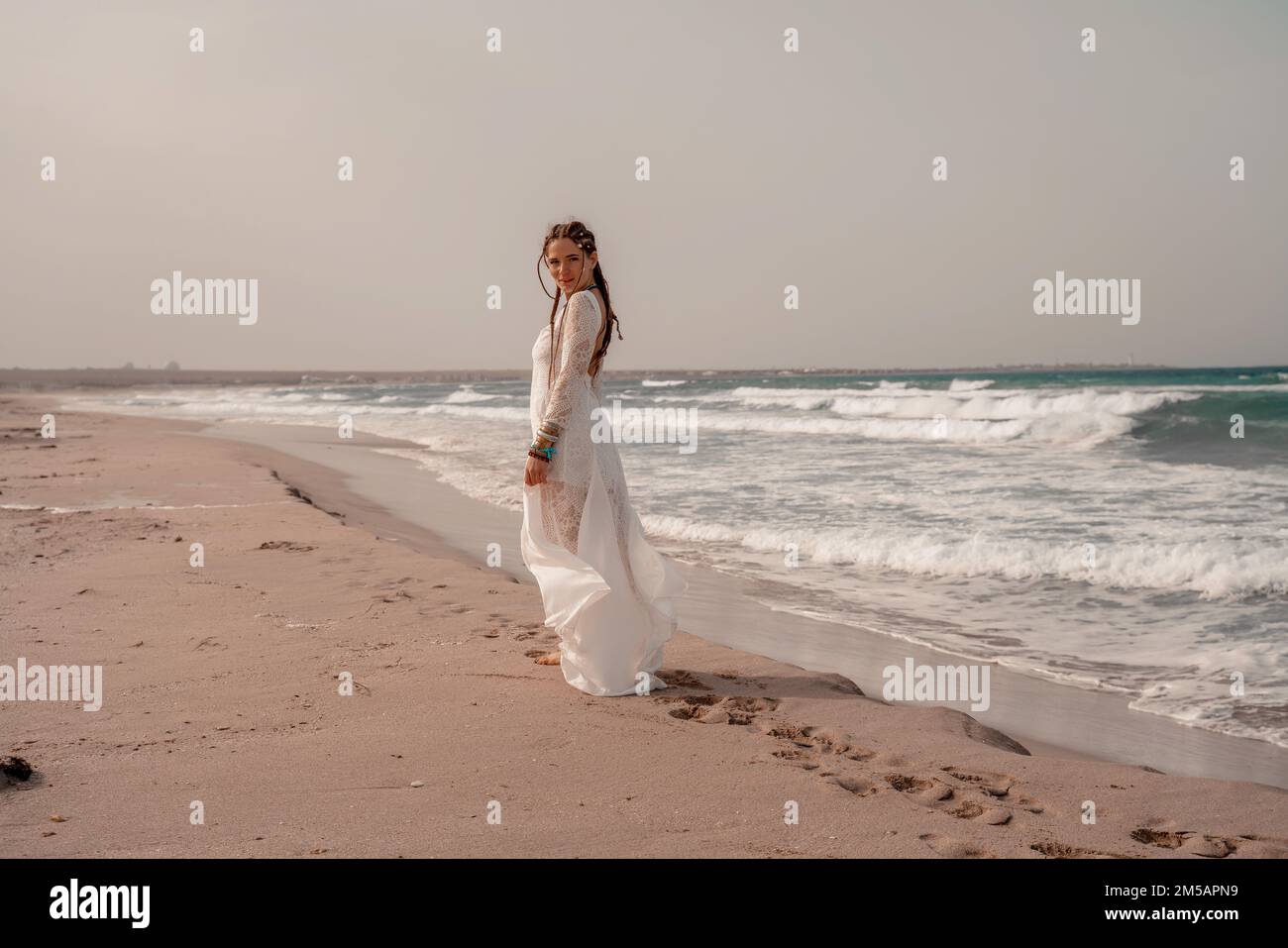 Model in boho style in a white long dress and silver jewelry on the beach. Her hair is braided, and there are many bracelets on her arms. Stock Photo