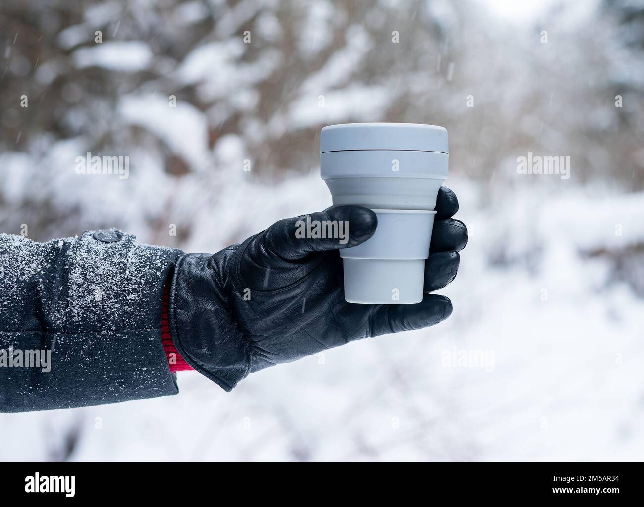 Hand in glove holding take away eco silicone coffee cup, take out tea mug in winter, in snowy weather outdoors. High quality photo Stock Photo