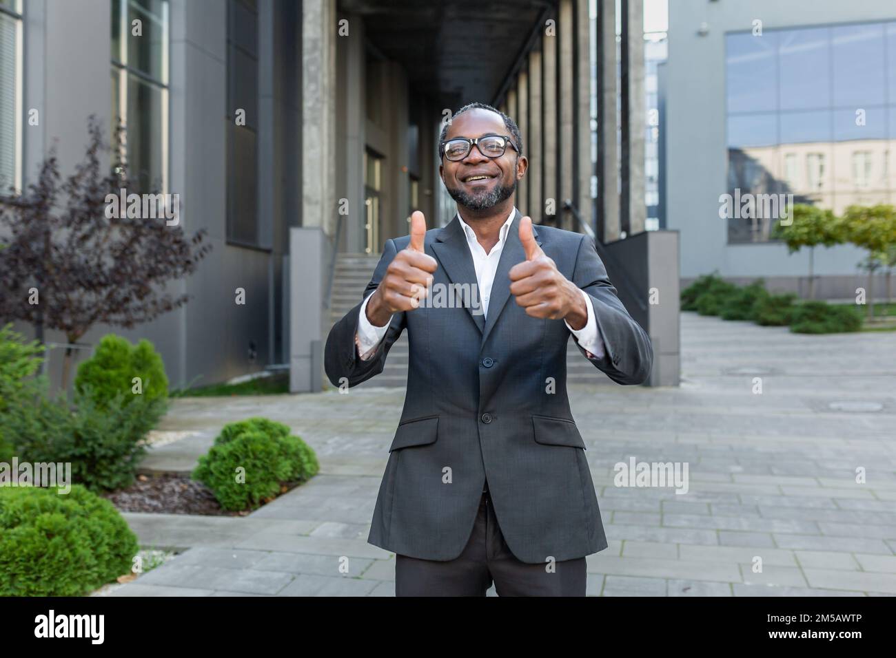 Portrait of successful African American mature businessman, senior man in business suit smiling and looking at camera showing happy thumbs up outside office building. Stock Photo