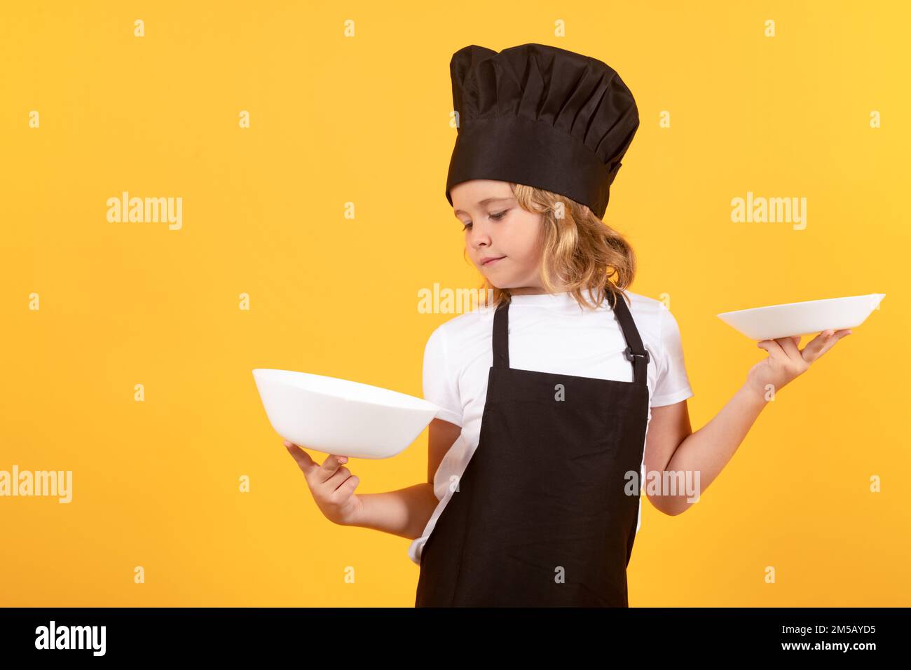 Little cook with cooking plate. Kid in cooker uniform and chef hat preparing food on studio color background. Cooking, culinary and kids food concept. Stock Photo