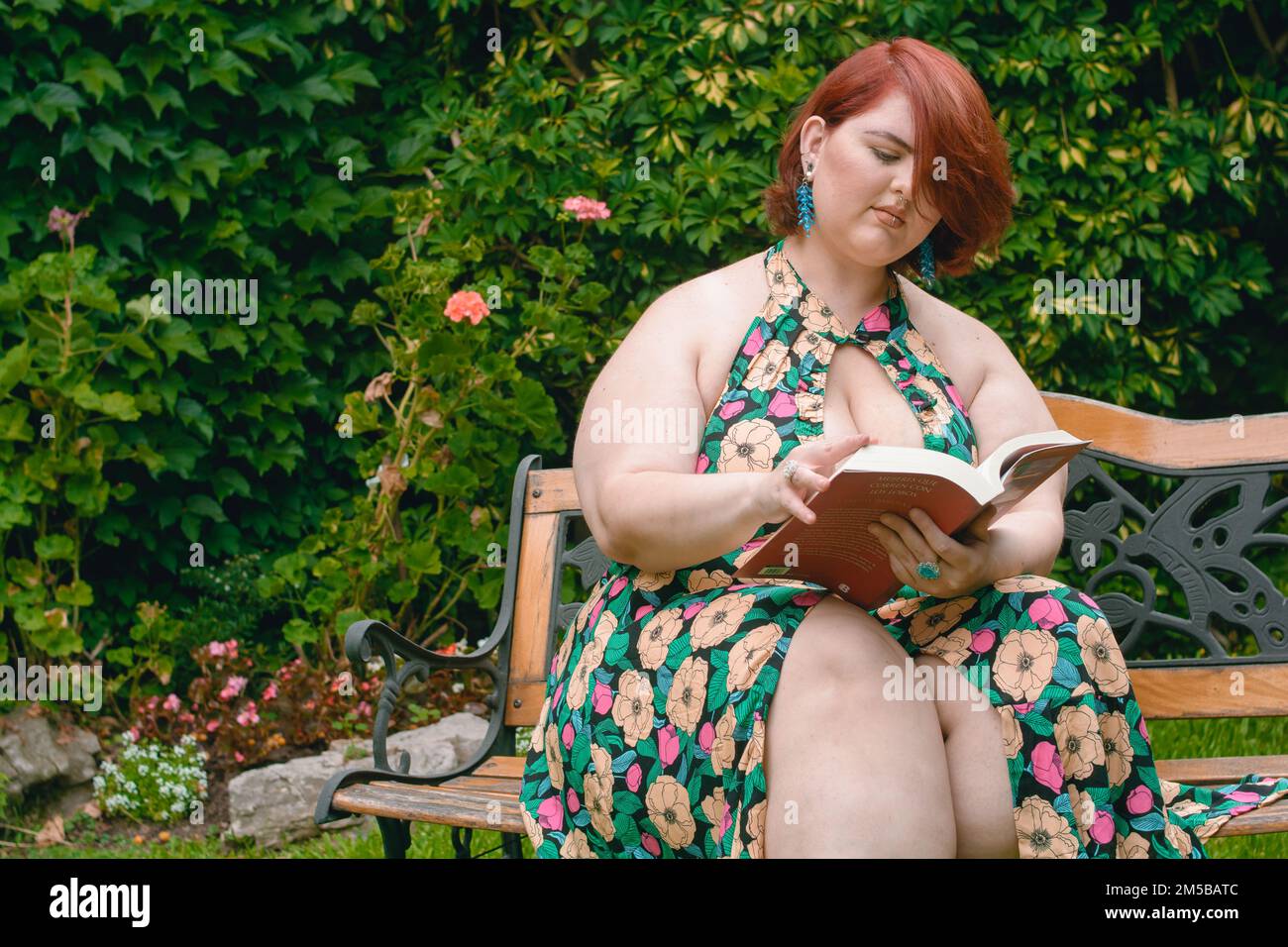 Caucasian Argentinian Latina woman plus size model reading a book sitting on a wooden bench in a garden, with a green wall in the background and copy Stock Photo