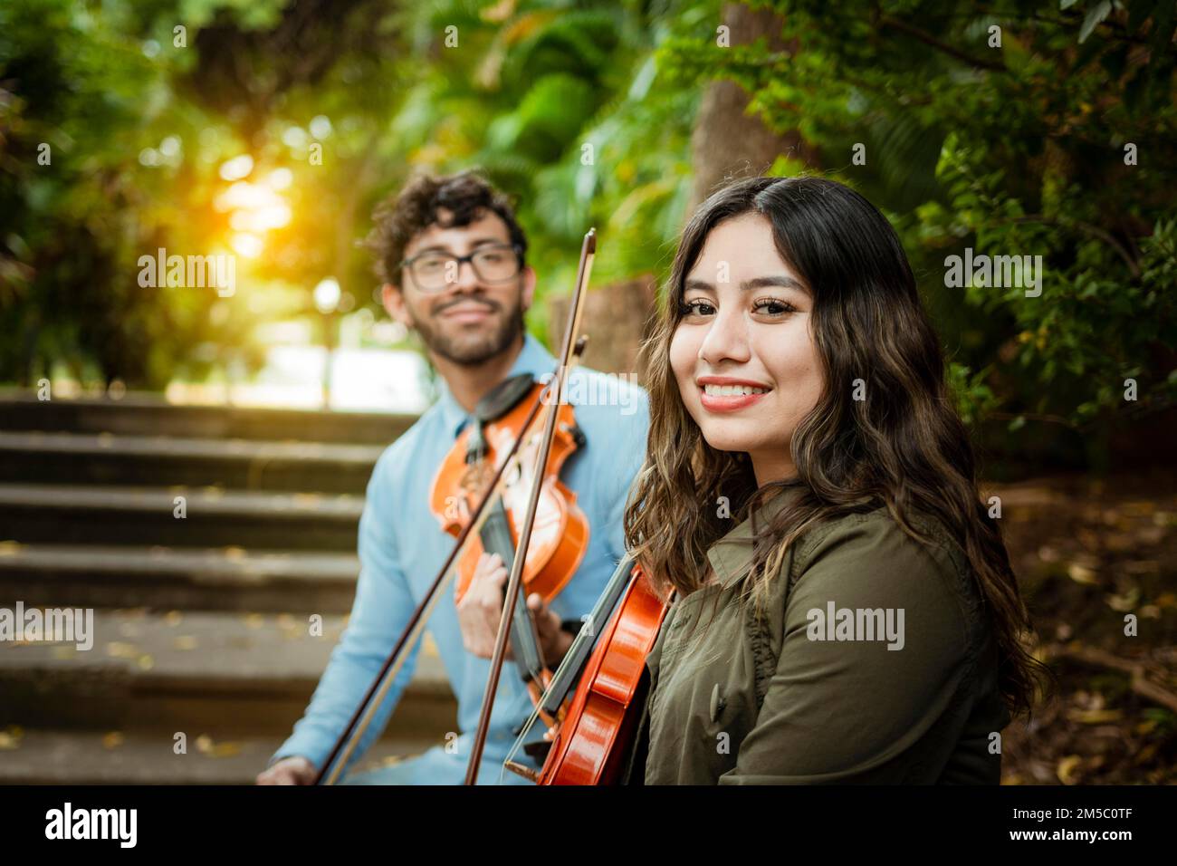 Two young violinists sitting outdoors looking at the camera. Portrait of two young violists sitting outside. Man and woman violinist sitting on the Stock Photo
