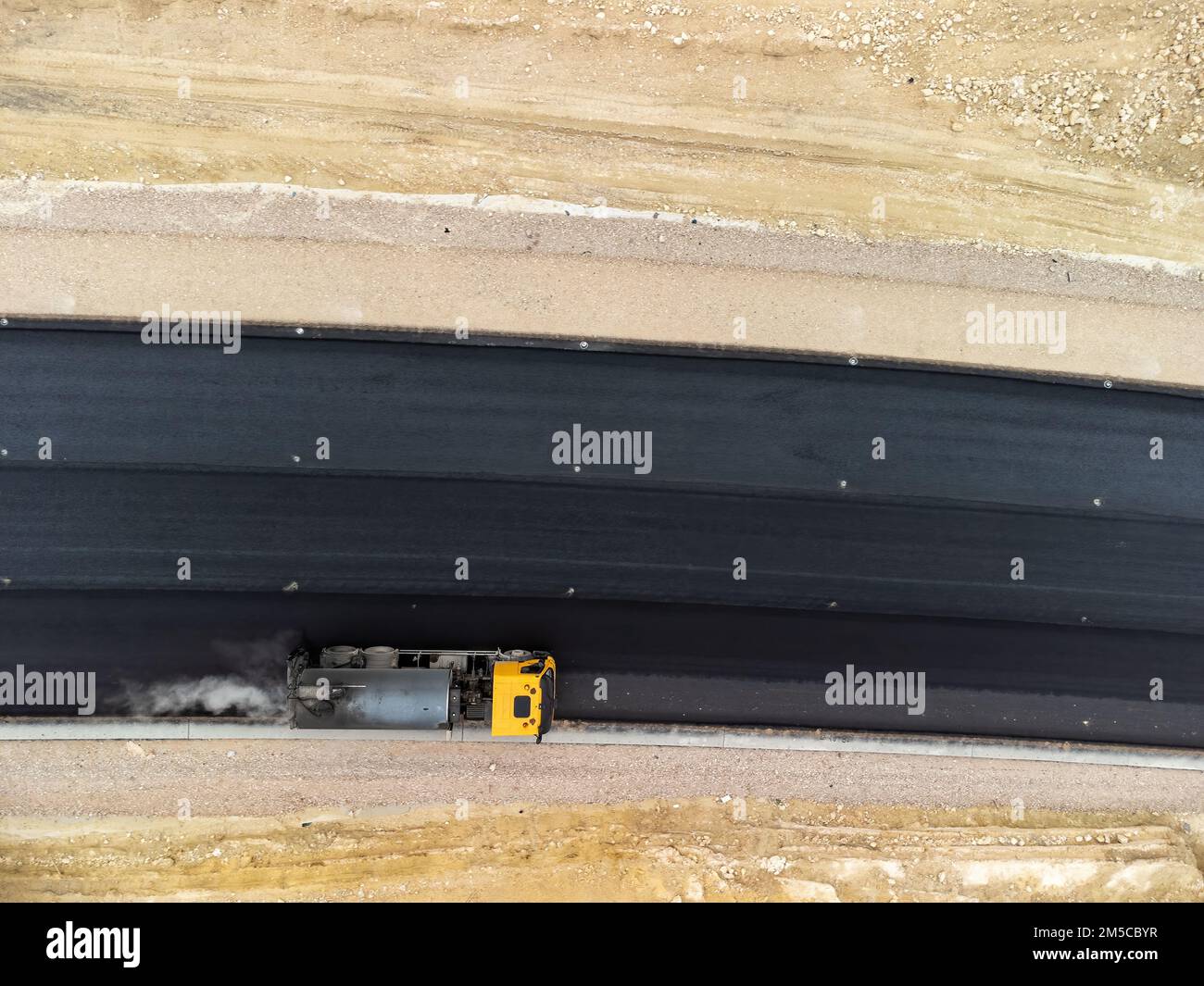 yellow truck pouring bitumen over a new road. Aerial drone shot. Road construction in progress on slope nature canyon. Infrastructure development and Stock Photo