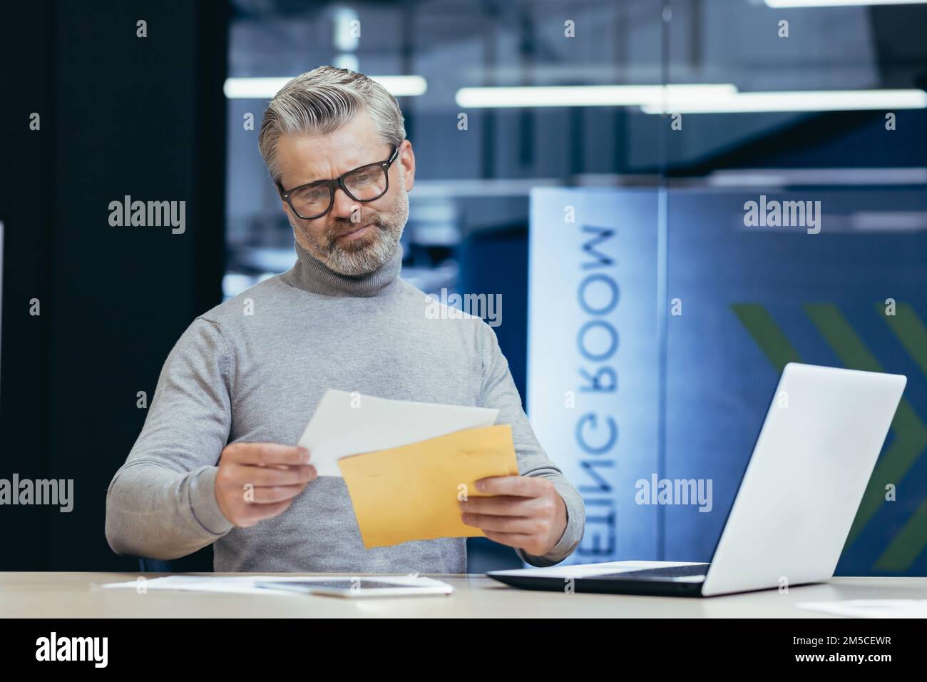 Mature gray-haired businessman inside modern office received letter message, senior man upset opens postal envelope, boss reads bad news upset and sad working at desk with laptop. Stock Photo