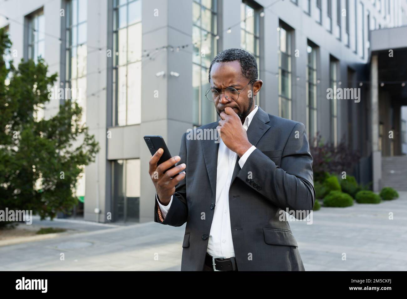 Mature senior african american boss reading bad news on phone, businessman in business suit outside office building using smartphone upset and disappointed by notification. Stock Photo