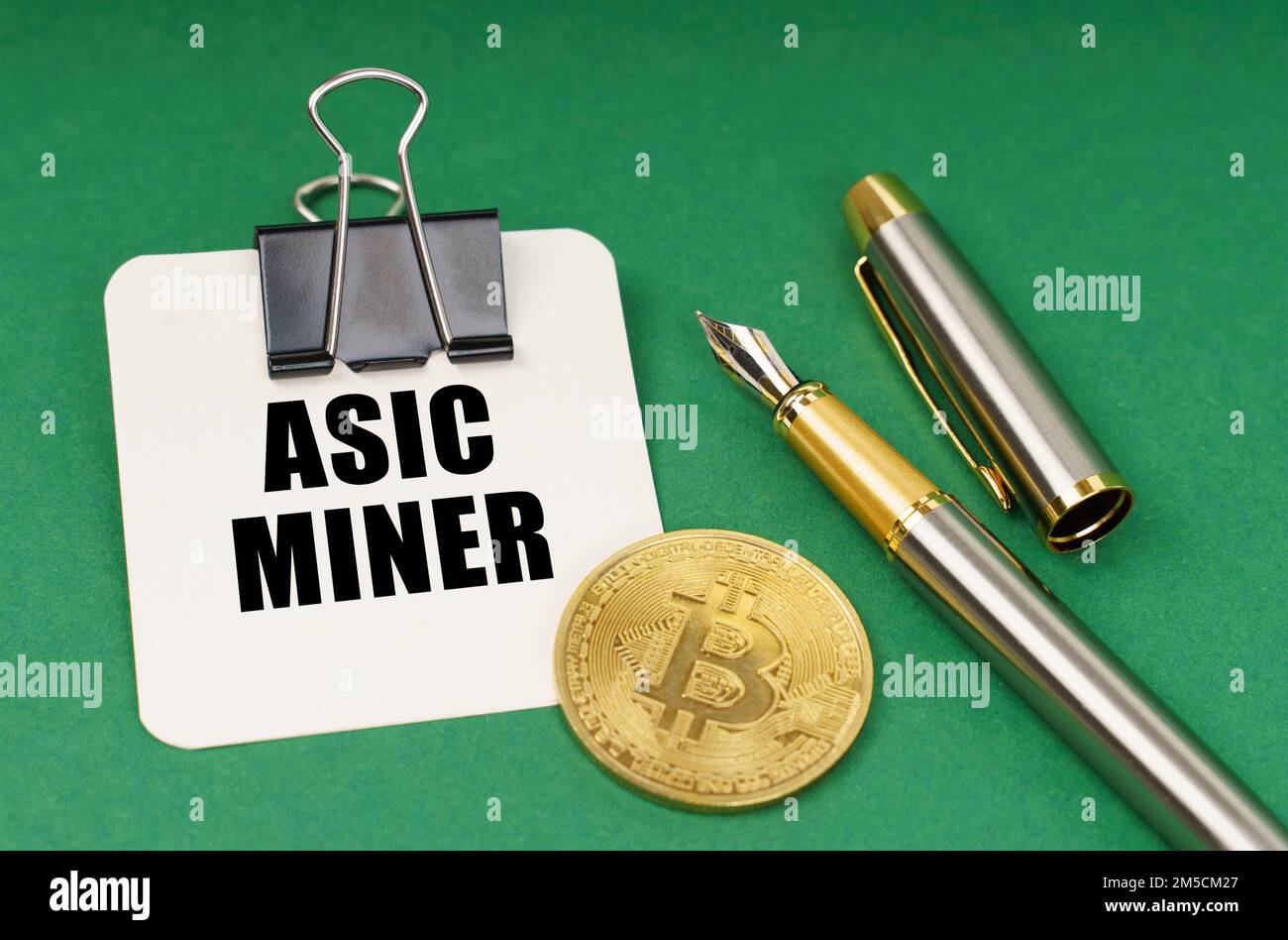 Cryptocurrency and business concept. On a green surface, a bitcoin coin, a pen and a sheet of paper with the inscription - Asic miner Stock Photo
