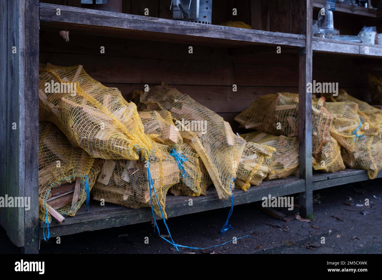 Small pieces of firewood, wrapped in a yellow nylon net, are displayed for sale in a metal rack. Easily combustible sticks or wood used to make fire Stock Photo