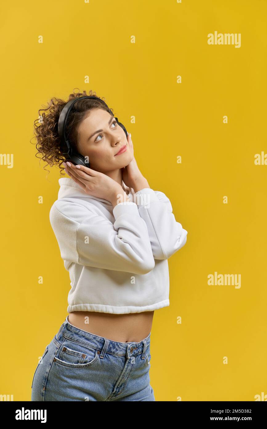 Curly haired woman listening music with wireless headphones in studio. Front view of cute pensive girl looking away, while enjoying music, isolated on orange studio background. Concept of relaxation. Stock Photo