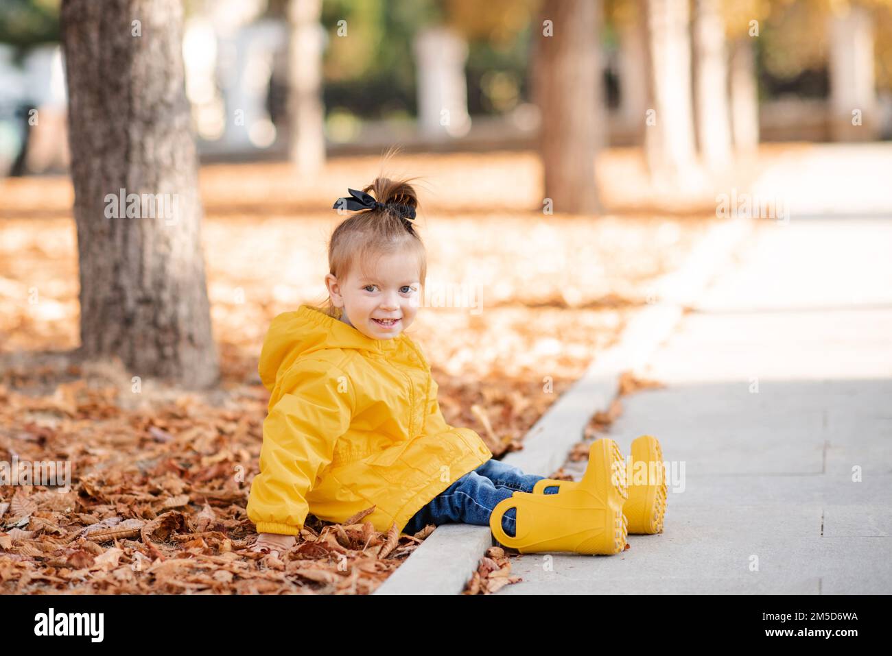 Cute funny kid girl 2-3 year old wear yellow rain jacket and rubber boots in autumn park outdoor over fallen leaves. Fall season. Childhood. Stock Photo