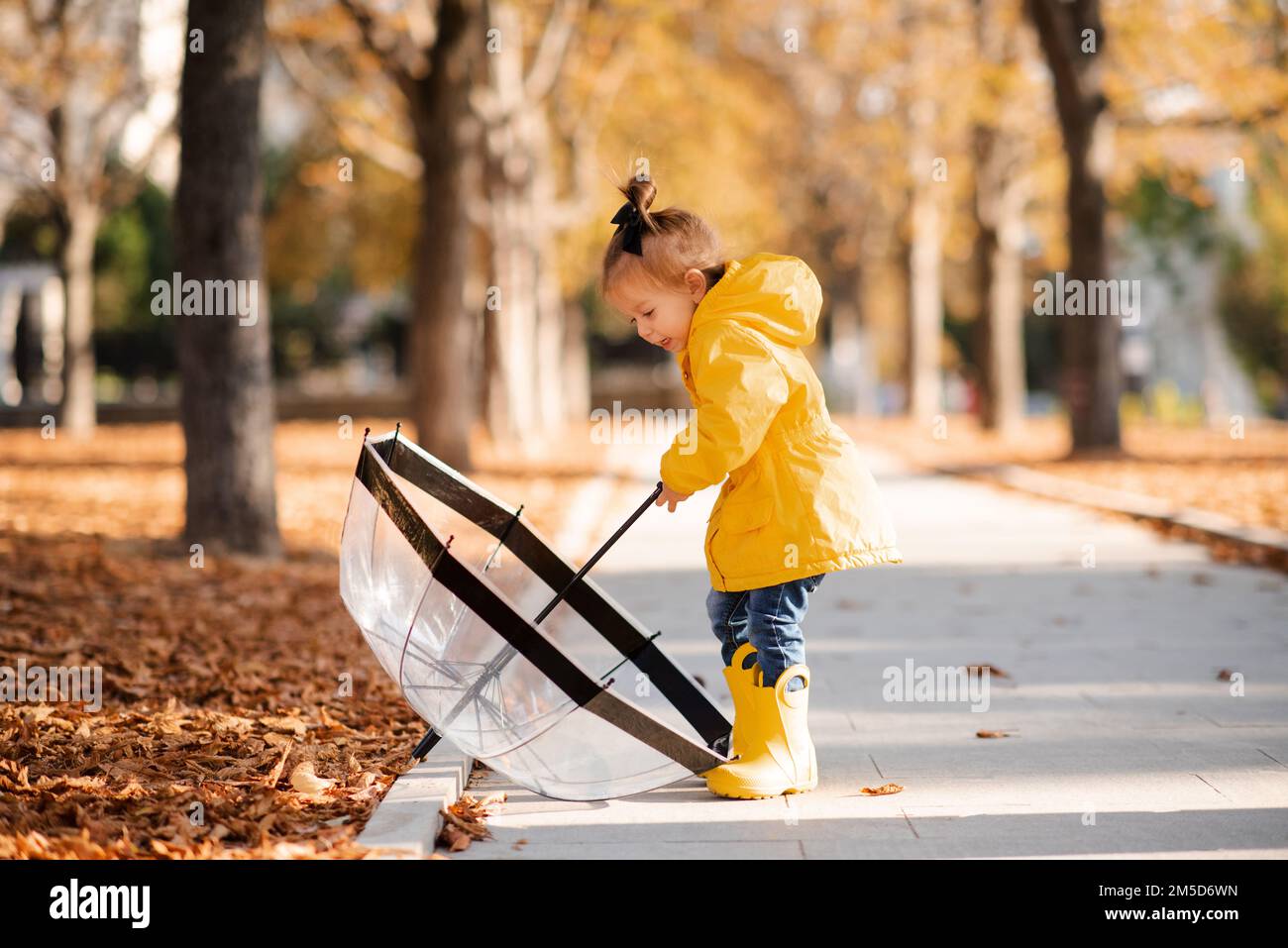 Cute funny kid girl 2-3 year old wear yellow rain jacket and boots hold umbrella in autumn park outdoor over fallen leaves. Fall season. Childhood. Stock Photo